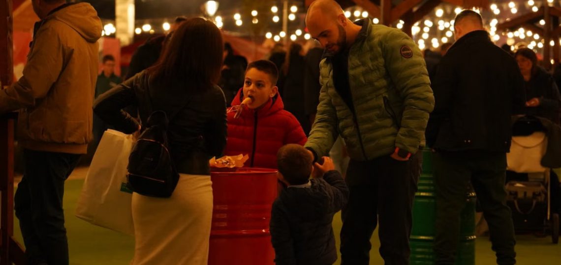 A family interacts at an evening street market with decorative lights and a bustling ambiance.